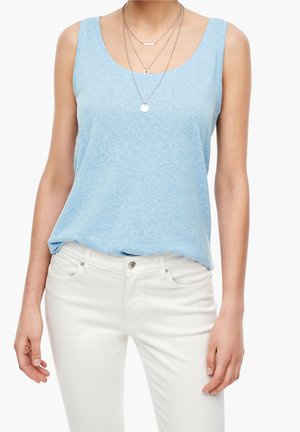 Kobieta ubrana w jasnoniebieską bluzkę bez rękawów, warstwowe srebrne naszyjniki i białe spodnie na jednolitym tle.