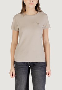 T-shirt beige a maniche corte con scollatura rotonda e un piccolo logo triangolare sul petto, abbinato a jeans grigi scuri. Tessuto dalla trama liscia.