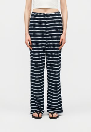 Žena oblečená v širokých nohaviciach s čierno-bielym cikcak vzorom, bielom krátkom topu a čiernych sandáloch, stojaca na jednoduchom pozadí.