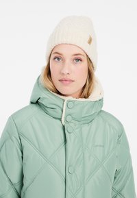 Hellgrüne, gesteppte Pufferjacke mit hohem Kragen, Knopfverschlüssen und cremefarbener Fleece-Fütterung, kombiniert mit einer cremefarbenen Strickmütze.