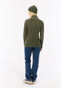 Olijfgroene geribde pullover met hoge kraag, gecombineerd met blauwe jeans en beige sneakers, van achteren gezien, met een gestructureerde stof.