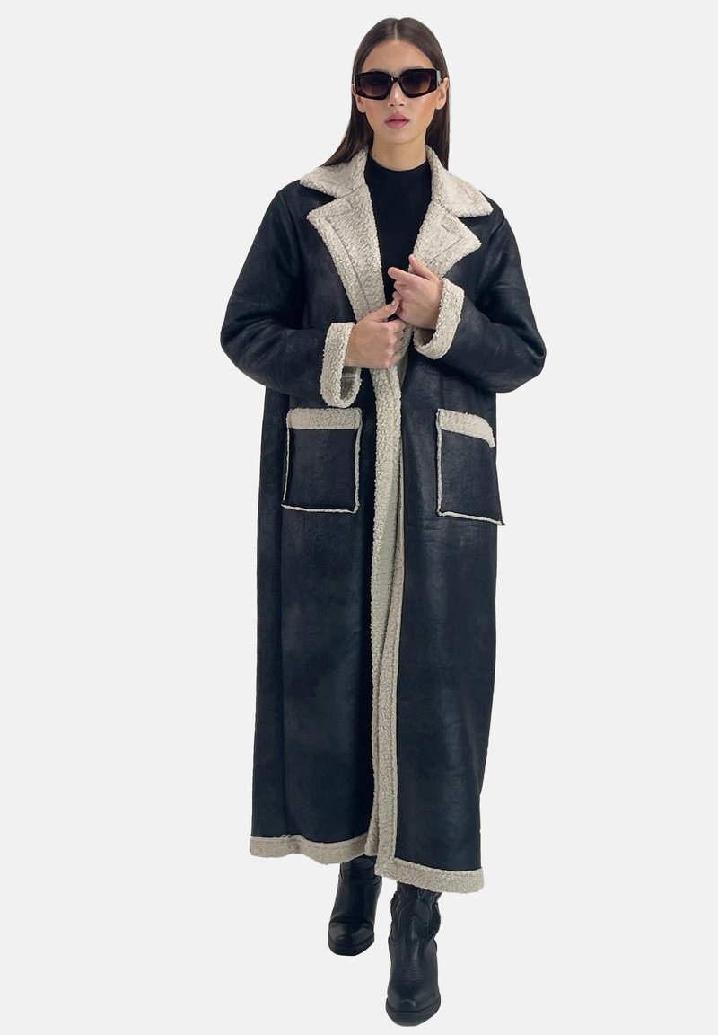 Abrigo largo negro con cuello y puños de piel sintética color crema, que cuenta con dos bolsillos delanteros y un diseño de corte recto. Se lleva sobre una blusa negra.