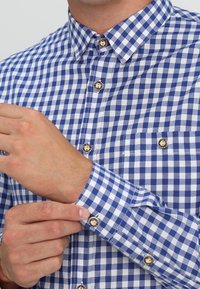 Mann passt den Bund seines blau-weißen karrierten Langarmhemds mit braunen Knöpfen an, das ein kreisförmiges schwarzes Design aufweist.