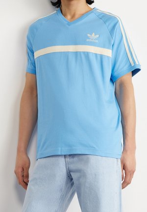 Personne portant un t-shirt Adidas bleu clair à col en V avec une bande horizontale blanche et un logo, associé à un jean bleu clair.