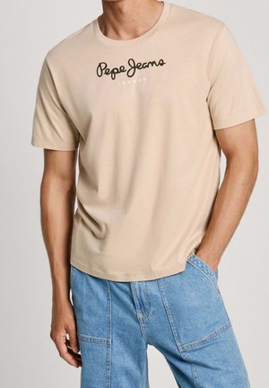 Beżowy t-shirt z krótkim rękawem z logo "Pepe Jeans London", noszony z jasnoniebieskimi spodniami dżinsowymi z naszywanymi kieszeniami.