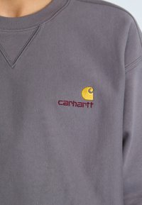 Siva dukserica s rebrastim izrezom, teksturiranom tkaninom i izvezenim žutim i burgundskim Carhartt logom na prsima.