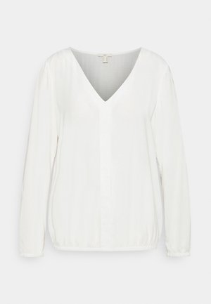 Blouse blanche à manches longues avec col en V, tissu doux, ourlet et poignets élastiques, présentée sur un fond clair uni.
