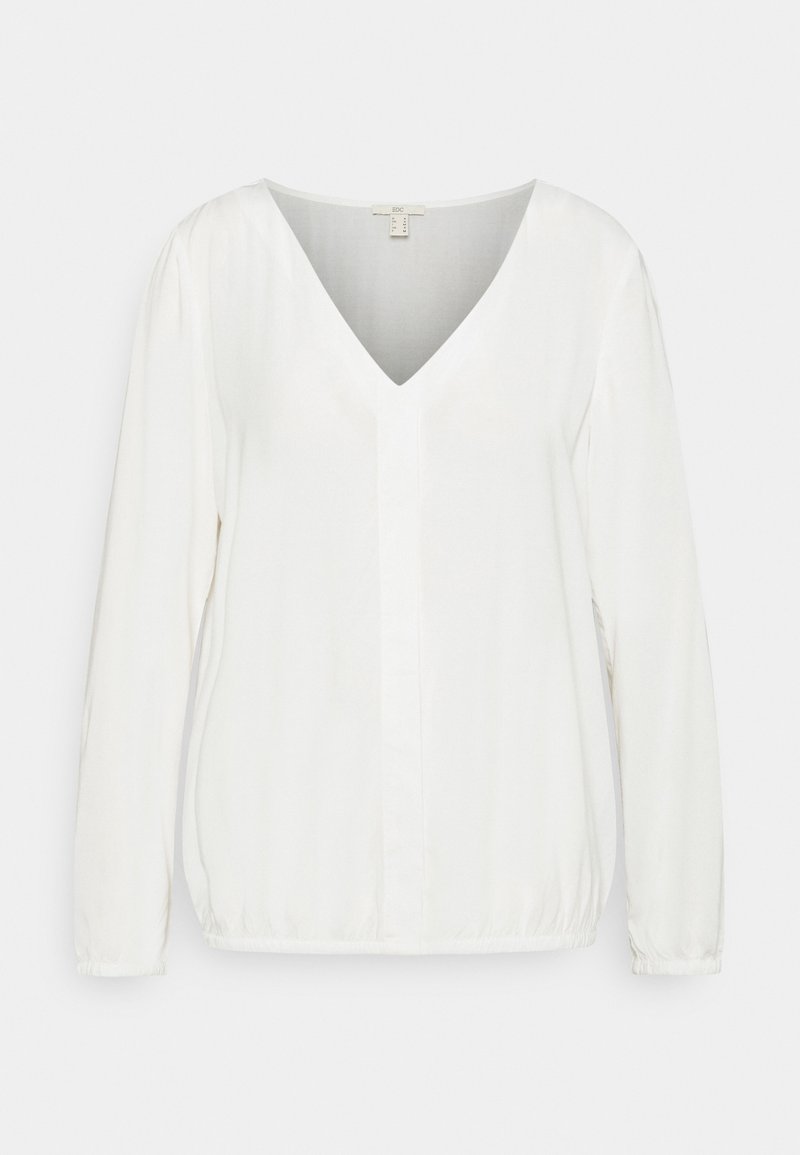 Blouse blanche à manches longues avec col en V, tissu doux, ourlet et poignets élastiques, présentée sur un fond clair uni.
