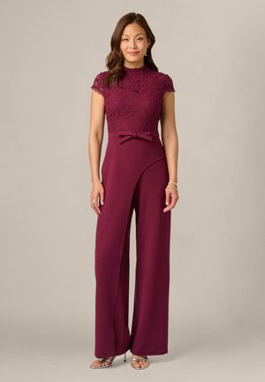 Femme debout portant une combinaison bordeaux sans manches avec un haut en dentelle, un col montant, un détail nœud à la taille, et un pantalon large avec des talons ouverts à l'avant.