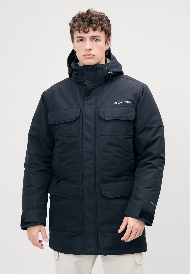 Zwarte geïsoleerde parka met een capuchon, meerdere voorzakken en een ritssluiting. Beschikt over een fleece-voering bij de kraag en verstelbare manchetten.