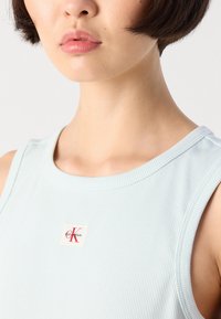 Svetlomodrý rebrovaný top bez rukávov s štvorcovou nášivkou s nápisom "Calvin Klein" v červenej farbe. Hladká textúra a okrúhly výstrih.