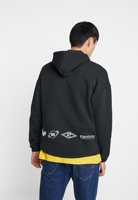 Svart huvtröja med avslappnad passform, texturerad tyg och logotypdetaljer på nederkanten av ryggen. Lager på en gul skjorta.