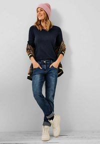 Marineblaue Langarmbluse, kariertes Jackett, blaue Jeans und cremefarbene Sneakers. Eine rosa gerippte Mütze setzt einen Farbakzent. Lässiges Outfit.