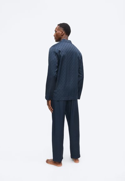 Férfi áll mezítláb, sötétkék mintás, hosszú ujjú felsőt és hozzá illő laza nadrágot visel, hátulról nézve, egyszerű világos háttér előtt.
