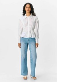 Witte blouse met lange mouwen en een getailleerde taille, knoopsluiting aan de voorkant en brede kraag, gecombineerd met lichtblauwe flared jeans. Platte sandalen maken de look compleet.