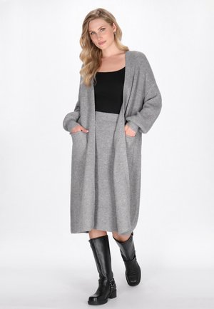 Lungo cardigan grigio con una texture a coste, maniche larghe e tasche laterali, indossato sopra una maglia nera e una gonna coordinata. Stivali neri al ginocchio.