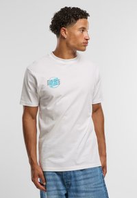 Hvit bomulls T-skjorte med rund hals, med en turkise sirkulært logo på venstre bryst og korte ermer. Denimshorts er på.