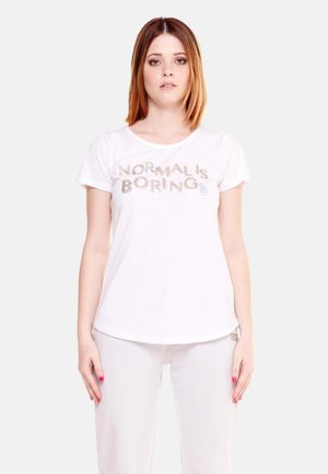 Giovane donna con capelli castano ramati lisci che indossa una T-shirt bianca con la scritta "NORMAL IS BORING" e pantaloni bianchi su uno sfondo semplice.