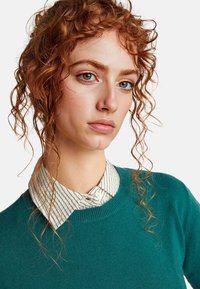 Teal pletený svetr s kulatým výstřihem, nošený přes bílou košili s modrými pruhy. Vlasy jsou stylizované do volných kudrn.