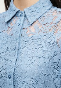 Chemise en dentelle bleue avec motifs floraux, devant boutonné et col, laissant voir la peau à travers le tissu en dentelle.