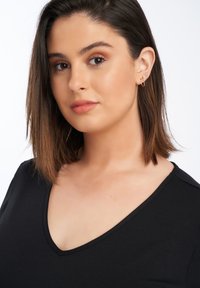 Zwarte V-hals top met een gladde textuur. Het model heeft schouderlang, steil bruin haar en draagt kleine hoopoorbellen.