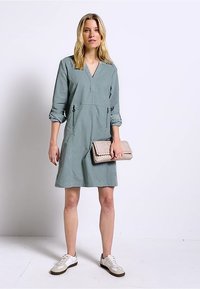 Ljusgrön knälång klänning med V-ringning, trekvartslånga ärmar och framfickor. Slät tyg kombinerat med en texturerad beige clutch.