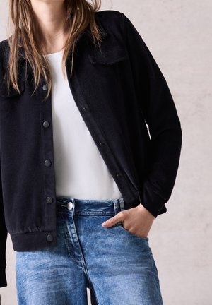 Femme portant une veste en velours côtelé noire boutonnée sur une chemise blanche et un jean bleu, main dans la poche, fond beige.