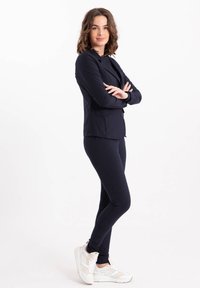 Marineblauwe fitted blazer en bijpassende legging, gecombineerd met witte sneakers. De blazer heeft een enkele knop en een gladde stofstructuur.