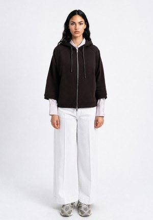Young woman stands wearing a black zip-up hoodie over a light striped shirt, white wide-leg pants, and gray sneakers on a plain background.