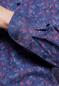 Koszula na guziki w kwiatowy wzór na granatowym tle, z małymi różowymi i niebieskimi kwiatkami. Mankiet ma rozcięcie zapinane na guzik.