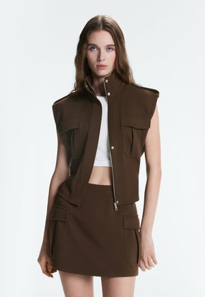 Frau trägt eine braune ärmellose Weste mit aufgesetzten Taschen vorne und einem passenden Rock mit Taschenklappen, kombiniert mit einem weißen, kurz geschnittenen Top.