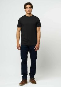 Vyras stovi, dėvėdamas paprastą juodą marškinėlį, tamsiai mėlynas džinsus ir rudos odos batus, priešais paprastą šviesią foną.