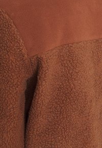 Hnědá fleecová látka se dvěma texturami: hladká horní část a měkká, kudrnatá spodní část s viditelným švem.