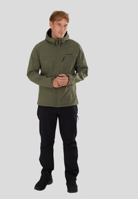 Oliven grøn softshell-jakke med en lynlåslomme på brystet, justerbar hætte og sorte lynlåsdetaljer, parret med sorte bukser og fodtøj.