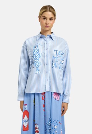 Woman wearing a blue and white striped shirt with fish and text "Take my dinner" and a blue skirt with food and bottle prints.