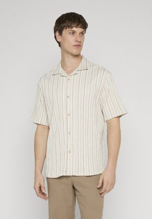 Młody mężczyzna w beżowej, krótkorękawowej koszuli na guziki z pionowymi paskami oraz jasnobrązowych spodniach, stojący na jednolitym jasnym tle.