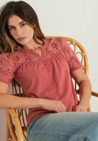 Top a maniche corte di colore rosa con motivi floreali ricamati, pattina con bottoni e vestibilità comoda, abbinato a jeans blu chiaro.
