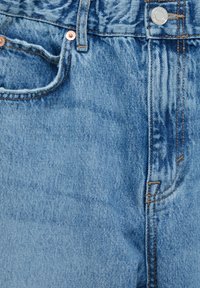 Gros plan sur un jean en denim bleu montrant la poche avant, la passants de ceinture, le bouton en métal et les rivets en cuivre avec la texture du tissu et les coutures visibles.