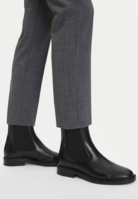 Czarne skórzane botki Chelsea z elastycznymi panelami bocznymi i gładkim wykończeniem, w zestawieniu ze szarymi, dopasowanymi spodniami.