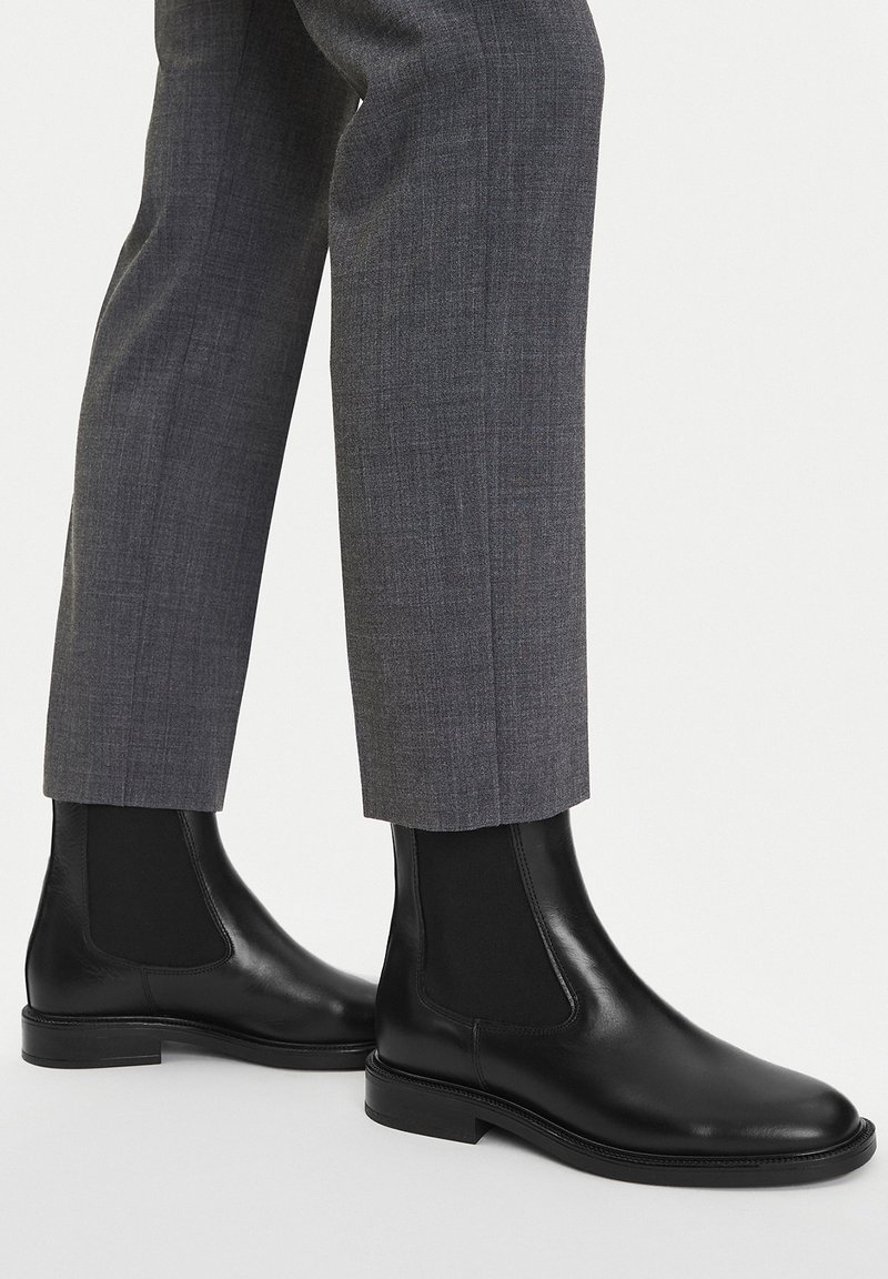 Czarne skórzane botki Chelsea z elastycznymi panelami bocznymi i gładkim wykończeniem, w zestawieniu ze szarymi, dopasowanymi spodniami.