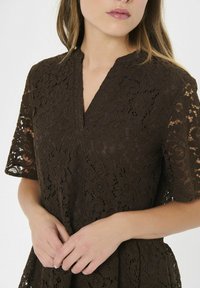 Naine kannab tumepruuni lillelise pitskleidi lühikeste varrukatega, V-kaelusega ja käed laisalt vööl.