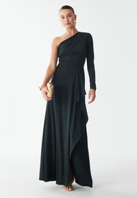Een schouderjurk in donkergroen met gedrapeerde details, gemaakt van zacht materiaal. Accentueert met een gouden clutch en sandalen met bandjes.