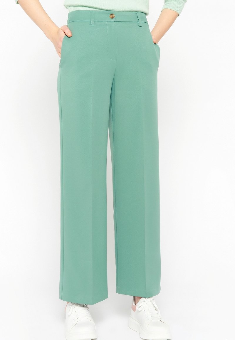 Femme portant un pantalon taille haute, jambes larges, vert menthe avec poches latérales et baskets blanches, debout devant un fond blanc uni.