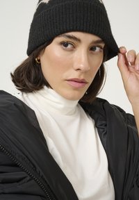 Bonnet en tricot noir, col roulé blanc, veste noire matelassée. Mise au point sur les traits du visage, peau lisse, cheveux foncés et boucle d'oreille discrète.