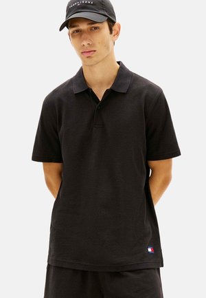 Junger Mann trägt schwarzes strukturiertes Poloshirt und passende Shorts, schwarze Tommy Jeans Cap, steht mit den Händen hinter dem Rücken.