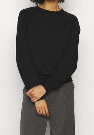 Persoon die een zwart trui met lange mouwen en een ronde hals draagt, samen met grijze broek, staand met de handen gevouwen voor zich tegen een effen achtergrond.