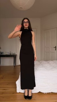 Νέα γυναίκα με μαύρο φόρεμα και γυαλιά που κουνάει το δεξί χέρι, στέκεται σε υπνοδωμάτιο με ξύλινο πάτωμα και άσπρο κρεβάτι.