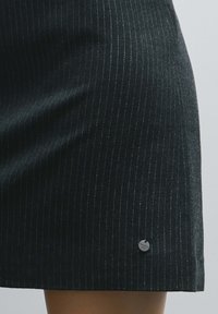 Gonna nera a righe sottili realizzata in tessuto strutturato, con un leggero scintillio e un piccolo dettaglio con logo metallico vicino all'orlo.