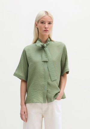 Vrouw met steil blond haar draagt een saliegroene blouse met korte mouwen en een strikkraag, gecombineerd met een witte broek, staand tegen een witte achtergrond.