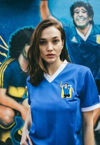 Camiseta de fútbol azul con cuello blanco, que presenta un gráfico de un jugador en el lado izquierdo del pecho. Tejido suave, mangas cortas y un corte relajado.